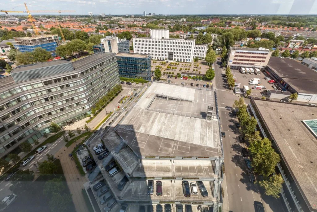 Luchtfoto van de Vliegend Hertlaan in een stedelijk gebied met kantoorgebouwen, een parkeergarage en woonwijken op de achtergrond.